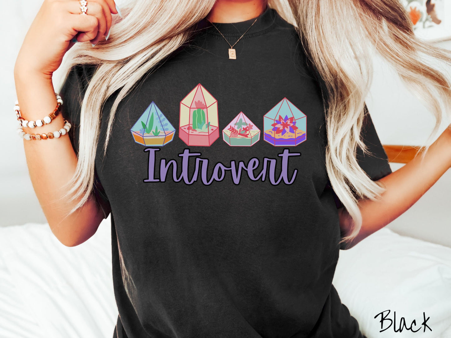 A woman wearing a cute, vintage black colored Comfort Colors t-shirt with the text Introvert in purple font across the front. Above the text are four colorful plant terrariums with colorful flowers and cacti inside.