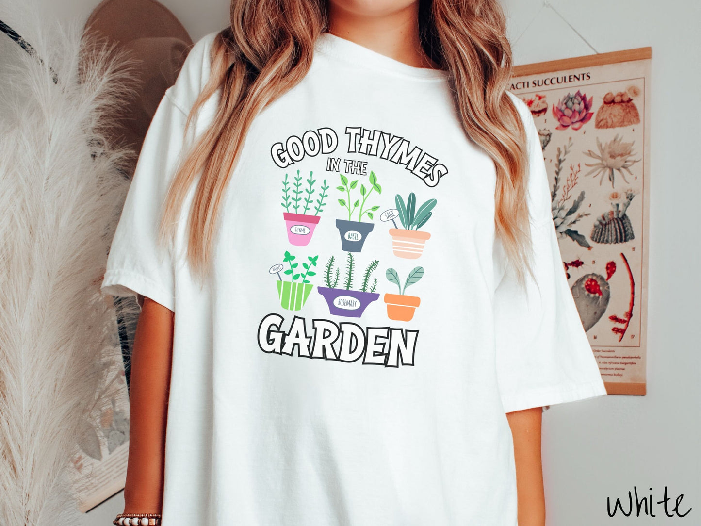 A woman wearing a cute, vintage white colored Comfort Colors t-shirt with the text Good Thymes in the Garden in white font. In between the text are different potted vegetables in colorful pots like thyme, basil, sage, and mint.