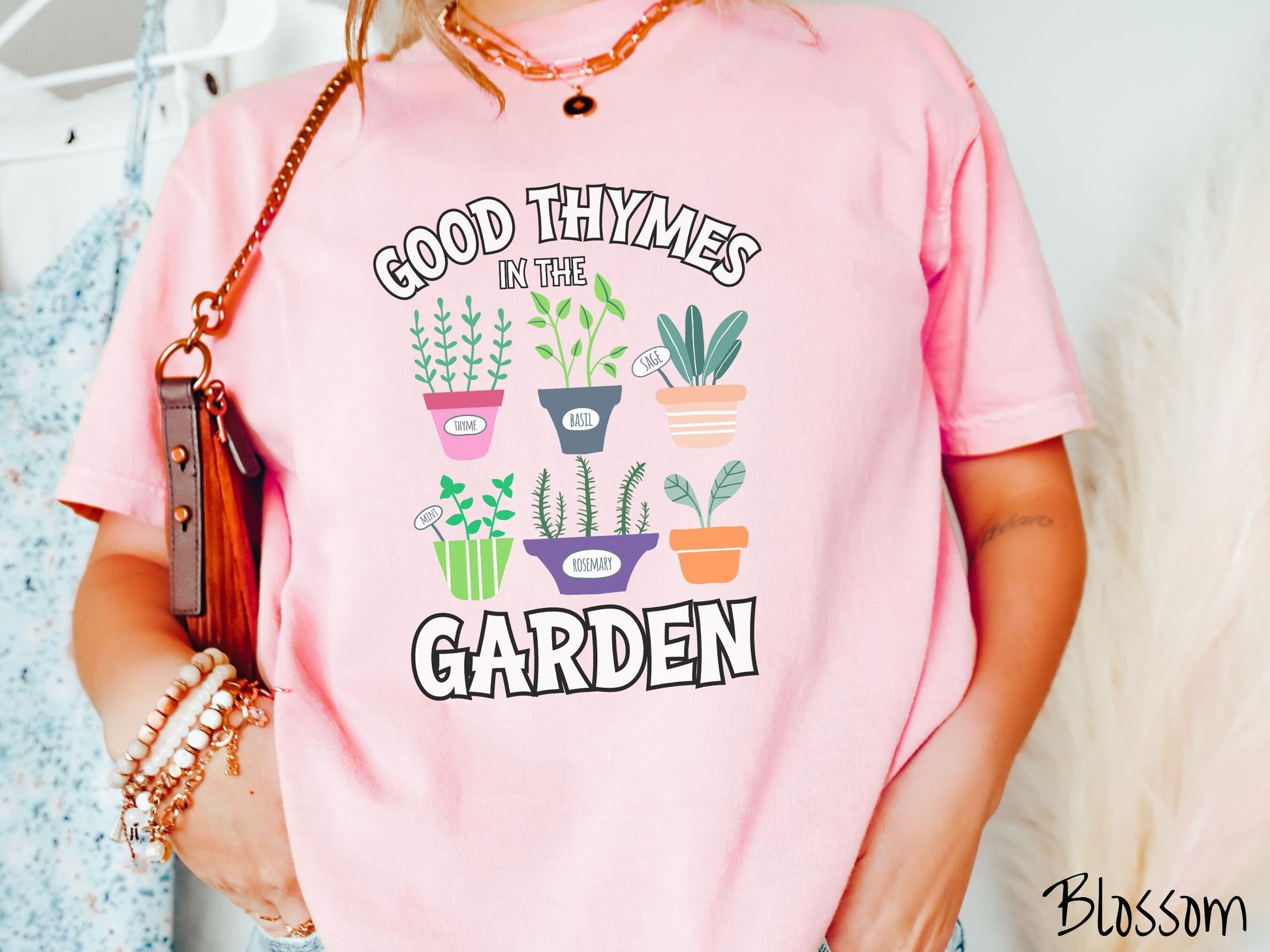 A woman wearing a cute, vintage blossom colored Comfort Colors t-shirt with the text Good Thymes in the Garden in white font. In between the text are different potted vegetables in colorful pots like thyme, basil, sage, and mint.