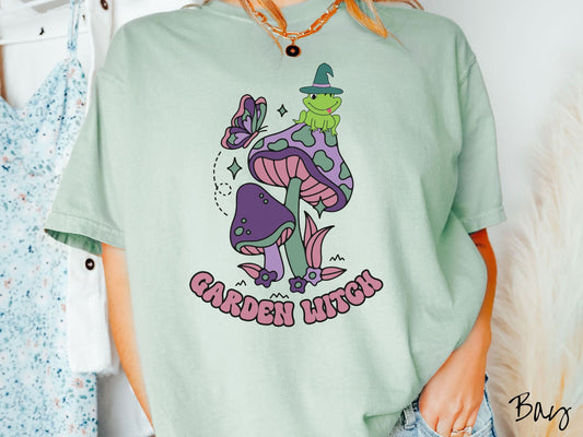 A woman wearing a cute, vintage bay colored Comfort Colors t-shirt with the text Garden Witch in purple font. Above this text are two purple and green mushrooms with a green frog sitting atop one wearing a witch hat.