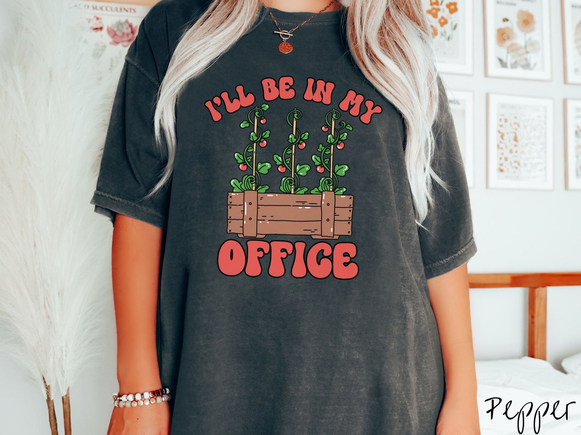 A woman wearing a cute, vintage pepper colored Comfort Colors t-shirt with the text I’ll Be in My Office in red font across the front. In between the text is a wooden plant holder with three tomato vines sprouting tomatoes climbing upwards.