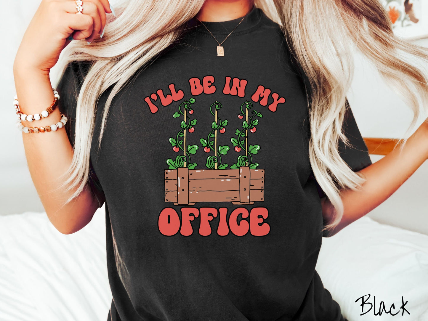 A woman wearing a cute, vintage black colored Comfort Colors t-shirt with the text I’ll Be in My Office in red font across the front. In between the text is a wooden plant holder with three tomato vines sprouting tomatoes climbing upwards.