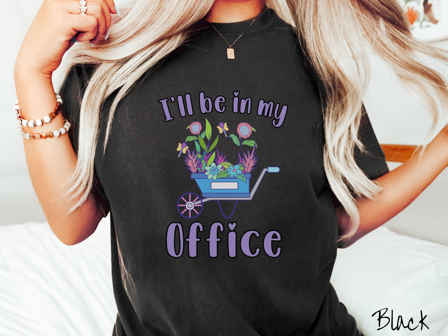 A woman wearing a cute, vintage black colored Comfort Colors t-shirt with the text I’ll Be in My Office in purple font across the front. In between the text is a blue wheelbarrow with a pink wheel stacked full of colorful plants and flowers.
