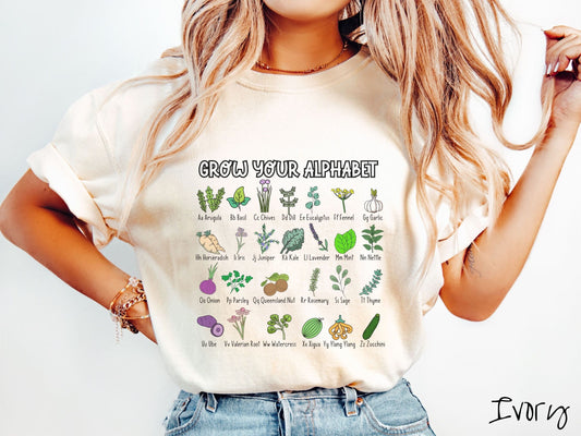 A woman wearing a cute, vintage ivory colored Comfort Colors t-shirt with the text Grow Your Alphabet across the top. Below this is a 4 x 7 grid of different kinds of plants and their names in alphabetical order  like Arugula, Basil, and Chives.