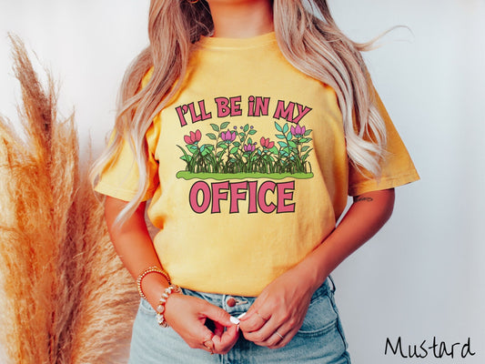 A woman wearing a cute, vintage mustard colored Comfort Colors t-shirt with the text I’ll Be in my Office in red font across the front. In between the text is a garden of red and pink flowers, green plants, and lush green grass.