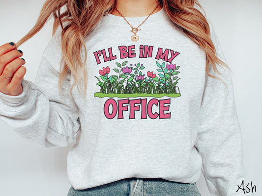 A woman wearing a cute, vintage ash colored comfy sweatshirt with the text I’ll Be in my Office in red font across the front. In between the text is a garden of red and pink flowers, green plants, and lush green grass.