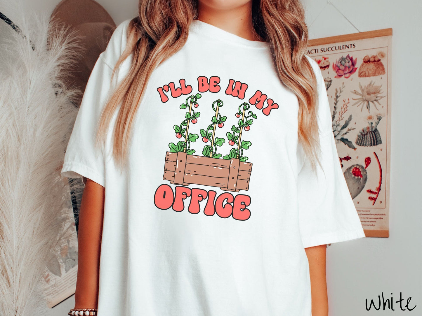 A woman wearing a cute, vintage white colored Comfort Colors t-shirt with the text I’ll Be in My Office in red font across the front. In between the text is a wooden plant holder with three tomato vines sprouting tomatoes climbing upwards.