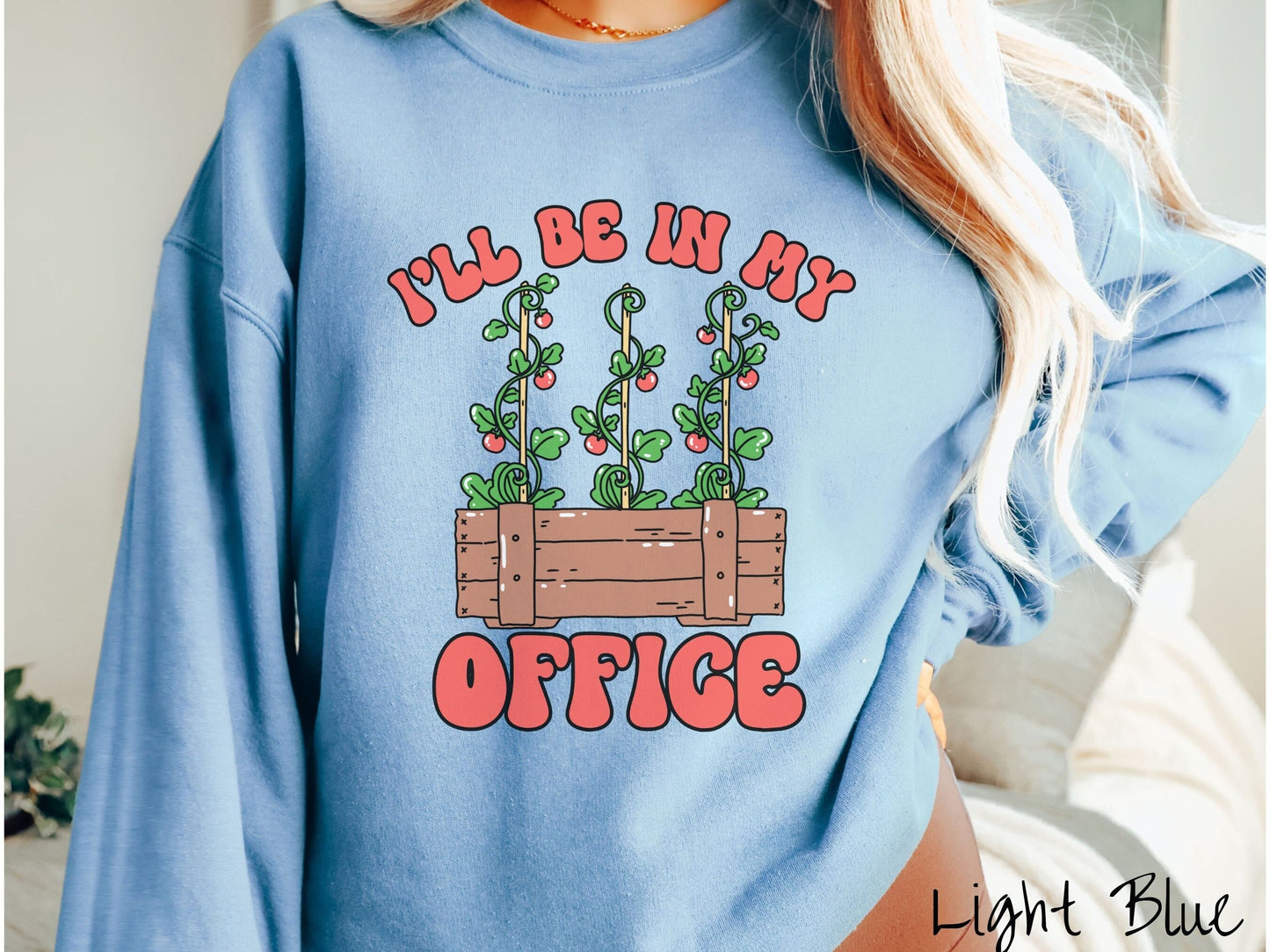 A woman wearing a cute, vintage light blue colored comfy sweatshirt with the text I’ll Be in My Office in red font across the front. In between the text is a wooden plant holder with three tomato vines sprouting tomatoes climbing upwards.