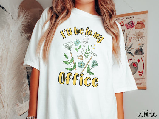 A woman wearing a cute, vintage white colored Comfort Colors t-shirt with the text I’ll Be in My Office in yellow font across the front. In between the text are gardening tools like a rake and shovel as well as different plants and flowers.