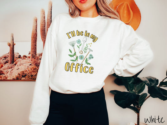 A woman wearing a cute, vintage white colored comfy sweatshirt with the text I’ll Be in My Office in yellow font across the front. In between the text are gardening tools like a rake and shovel as well as different plants and flowers.