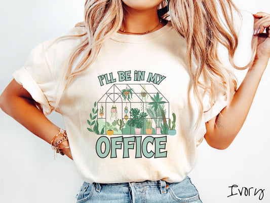 A woman wearing a cute, vintage ivory colored Comfort Colors t-shirt with the text Ill Be in My Office in green font across the front. In between the text is a glass greenhouse with all kinds of plants and flowers inside and hanging potted plants.