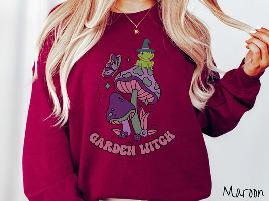 A woman wearing a cute, vintage maroon colored comfy sweatshirt with the text Garden Witch in purple font. Above this text are two purple and green mushrooms with a green frog sitting atop one wearing a witch hat.