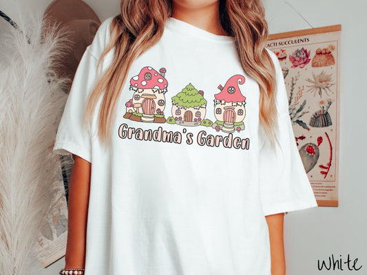 A woman wearing a cute, vintage white colored Comfort Colors t-shirt with the text Grandmas Garden in light pink font. Above the text are cute little cottages for gnomes made from light and dark pink mushrooms.