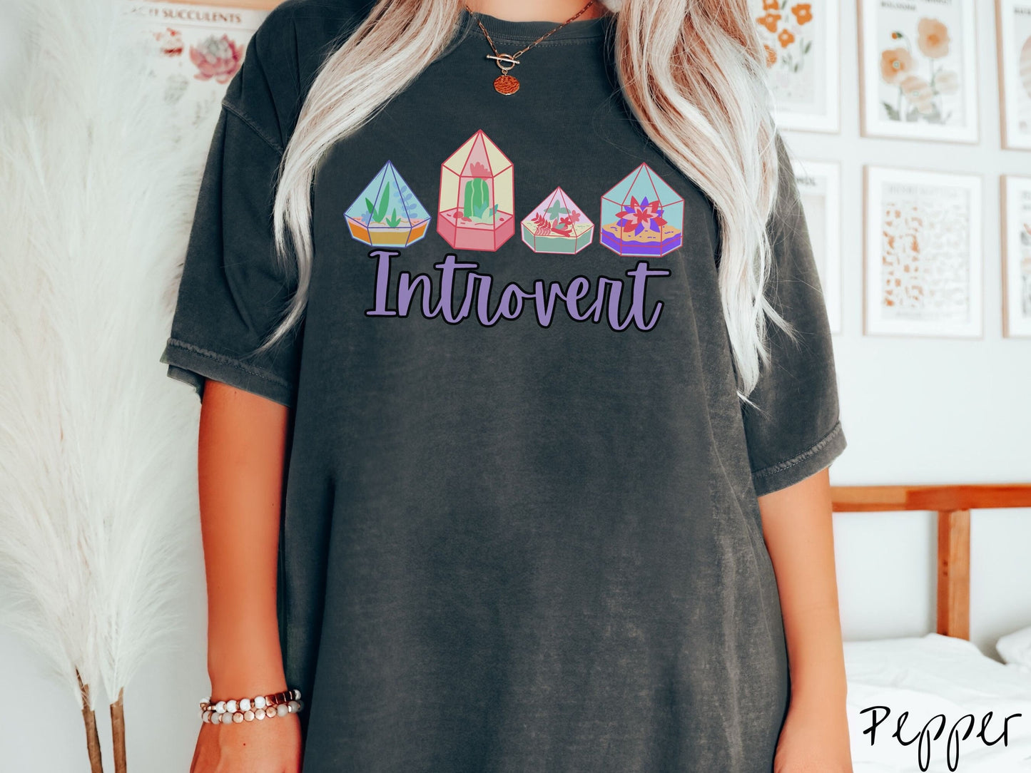 A woman wearing a cute, vintage pepper colored Comfort Colors t-shirt with the text Introvert in purple font across the front. Above the text are four colorful plant terrariums with colorful flowers and cacti inside.