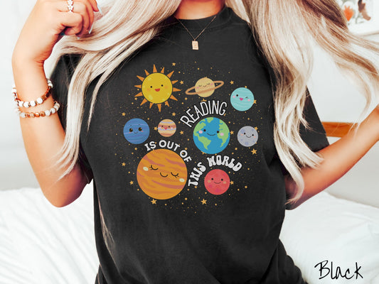 A woman wearing a cute, vintage black colored shirt showing the solar system and all the planets have smiling, happy faces. There is text saying Reading Is Out of This World among the planets, and there are many stars twinkling in the background.