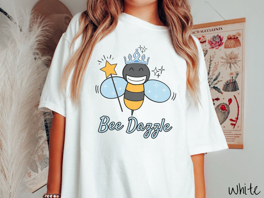 A woman wearing a cute, vintage white colored Comfort Colors t-shirt with text Bee Dazzle in light blue, cursive font. Above is a black and yellow bee with a wide smile and blue wings. It’s holding a star-tipped wand and wearing a light blue tiara.