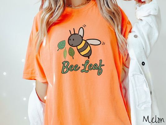 A woman wearing a cute, vintage melon colored Comfort Colors t-shirt with the text Bee Leaf in green, cursive font. Above that is a black and yellow honey bee buzzing through the air with green leaves floating next to it.