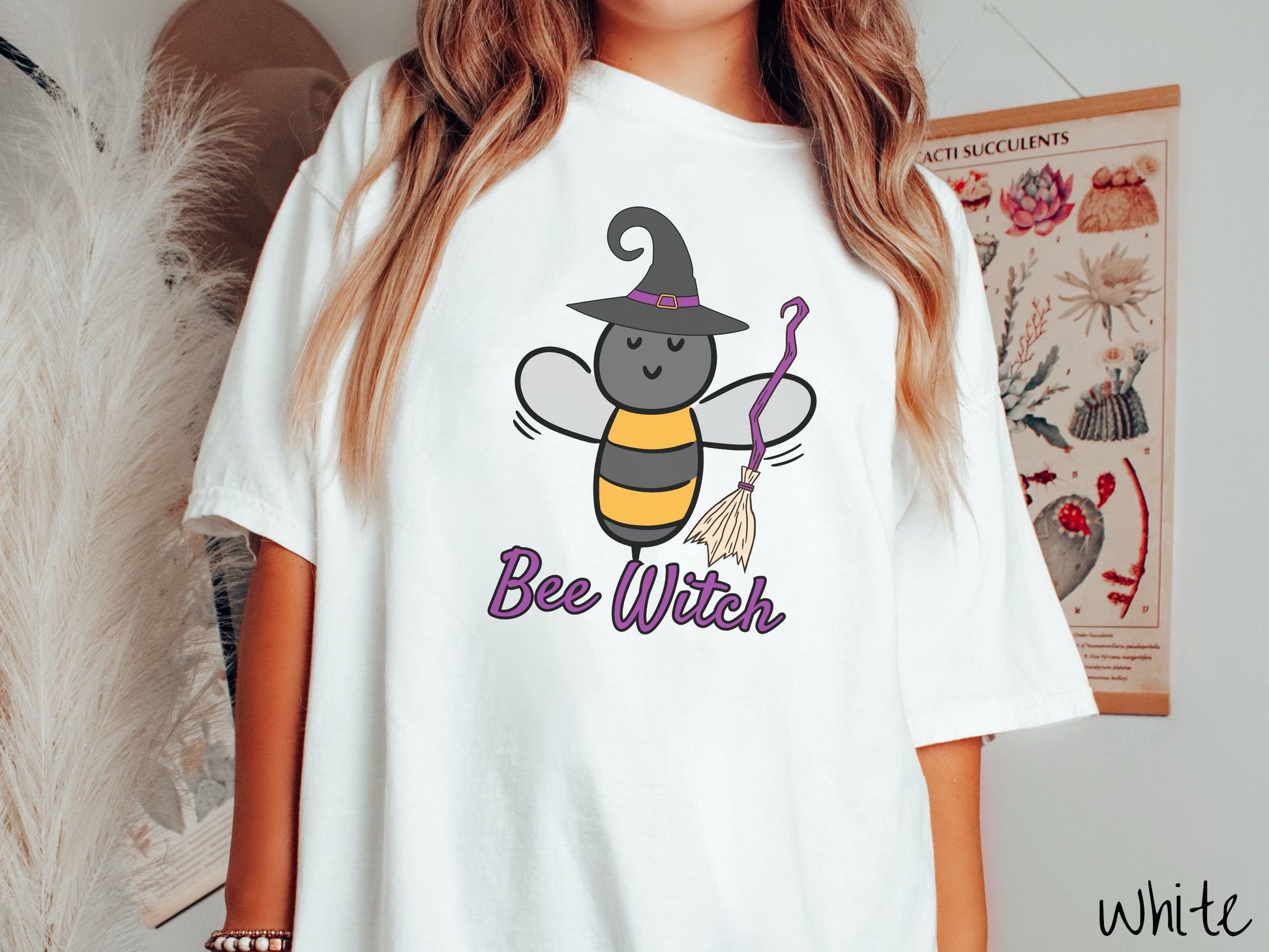 A woman wearing a cute, vintage white colored Comfort Colors t-shirt with the text Bee Witch in purple, cursive font. Above that is a black and yellow honey bee holding a purple, straw broom and wearing a black witch hat with a purple hat band.