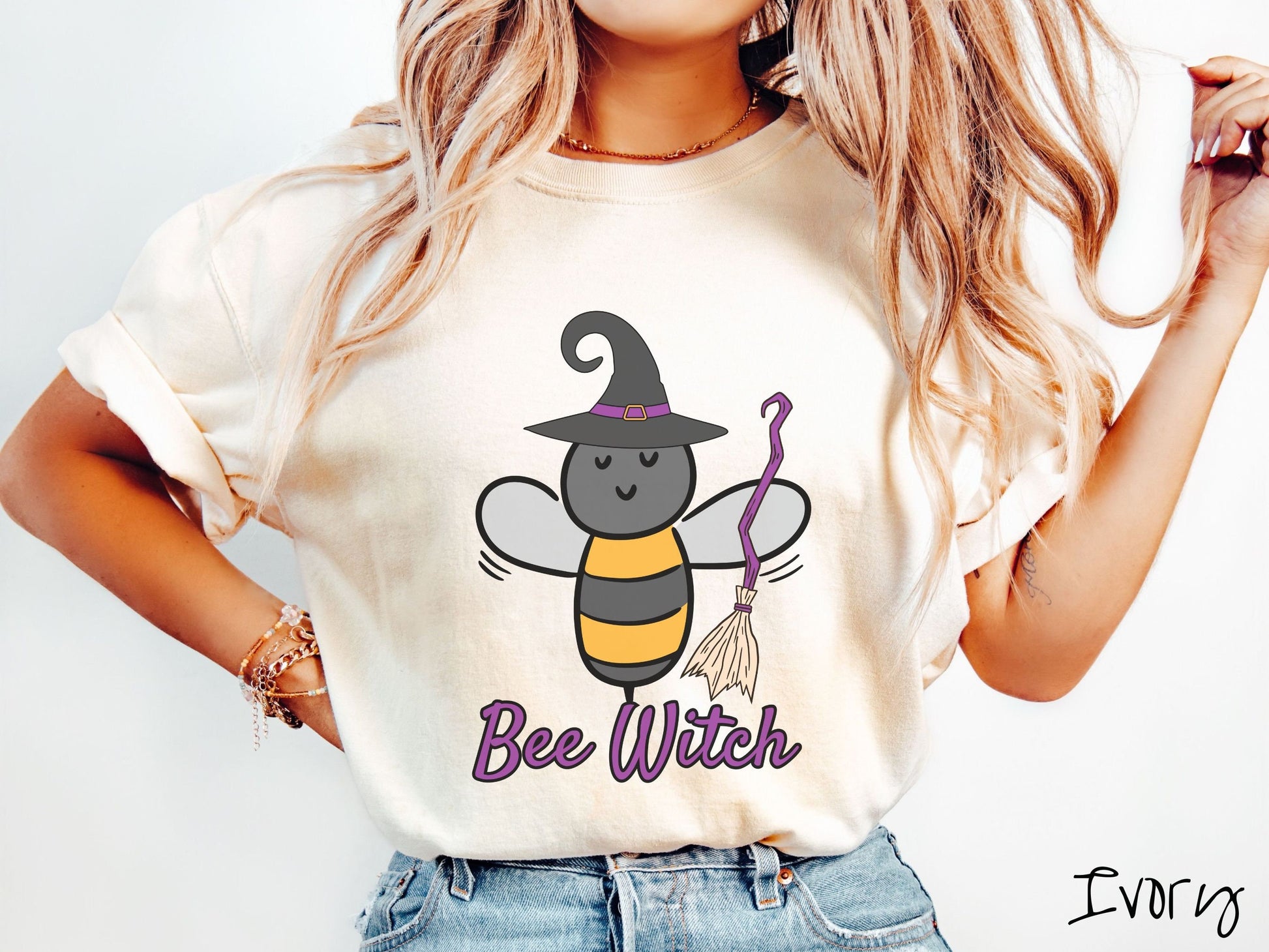 A woman wearing a cute, vintage ivory colored Comfort Colors t-shirt with the text Bee Witch in purple, cursive font. Above that is a black and yellow honey bee holding a purple, straw broom and wearing a black witch hat with a purple hat band.