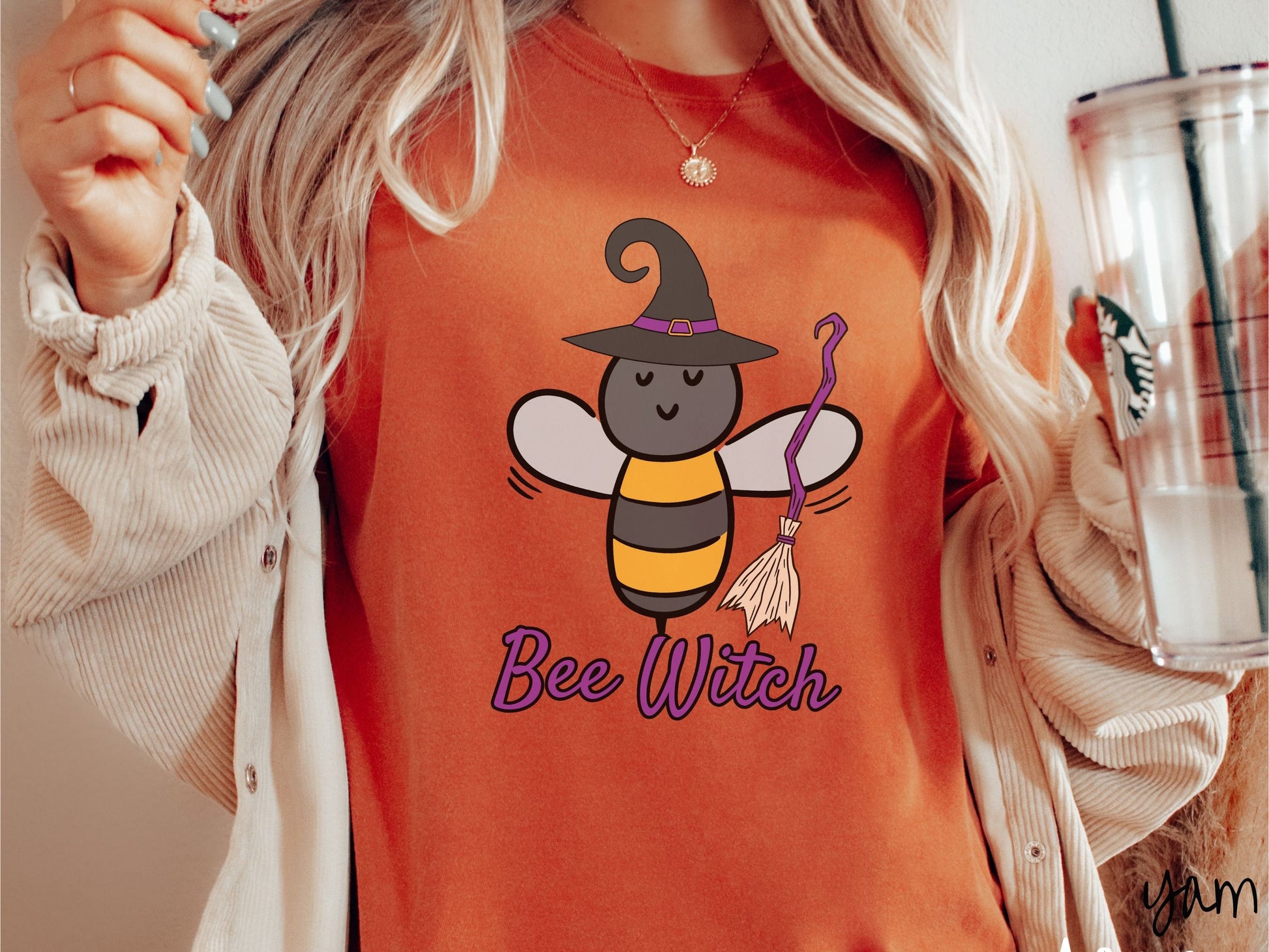 A woman wearing a cute, vintage yam colored Comfort Colors t-shirt with the text Bee Witch in purple, cursive font. Above that is a black and yellow honey bee holding a purple, straw broom and wearing a black witch hat with a purple hat band.