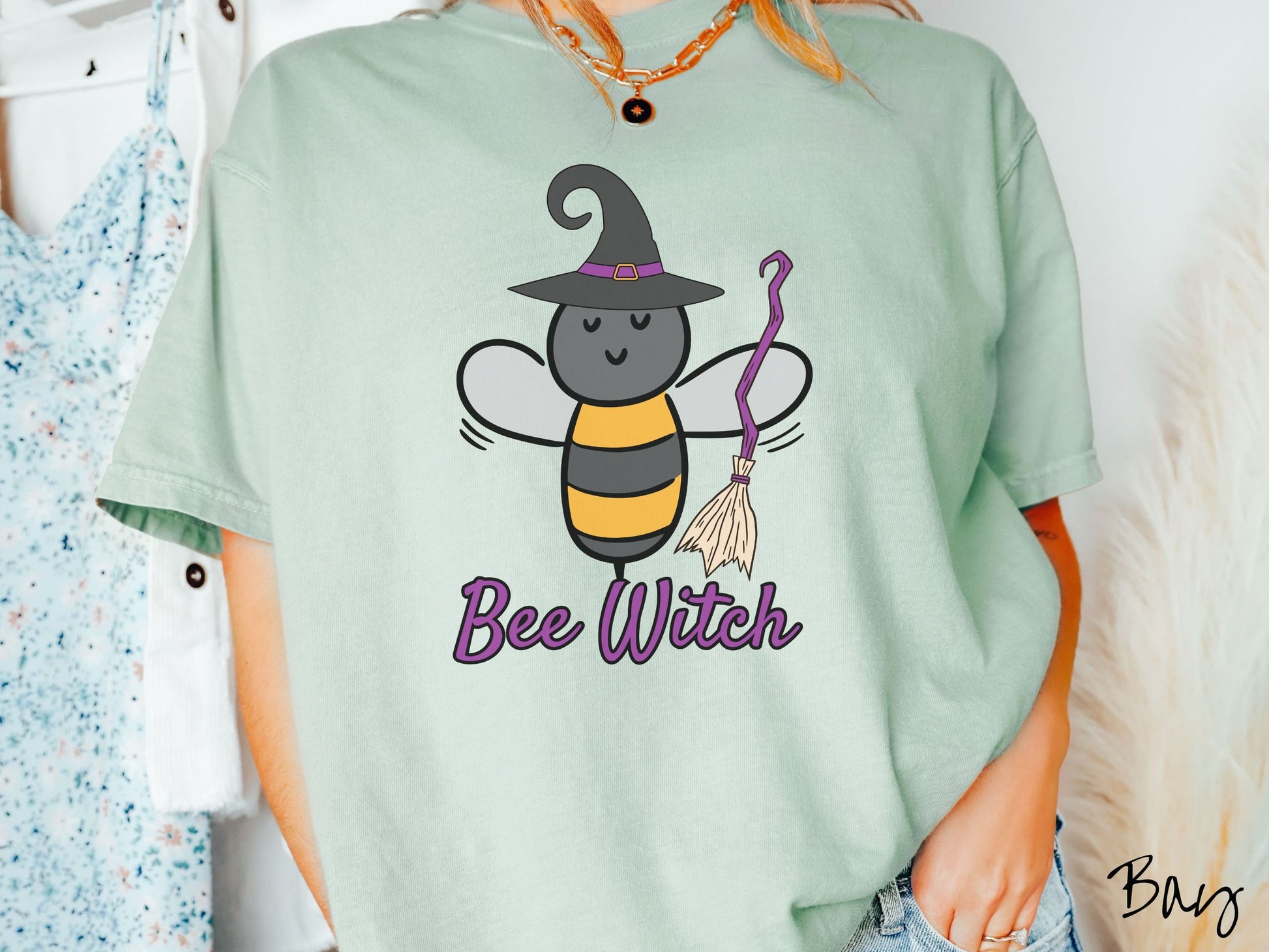 A woman wearing a cute, vintage bay colored Comfort Colors t-shirt with the text Bee Witch in purple, cursive font. Above that is a black and yellow honey bee holding a purple, straw broom and wearing a black witch hat with a purple hat band.