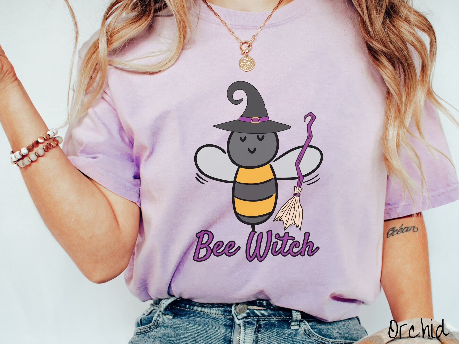 A woman wearing a cute, vintage orchid colored Comfort Colors t-shirt with the text Bee Witch in purple, cursive font. Above that is a black and yellow honey bee holding a purple, straw broom and wearing a black witch hat with a purple hat band.