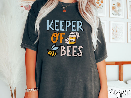 A woman wearing a cute, vintage pepper colored Comfort Colors t-shirt with the text Keeper of Bees in blue, orange, and pink uppercase font. In between the text is a yellow pot of honey and a black and yellow bee with blue wings buzzing around.