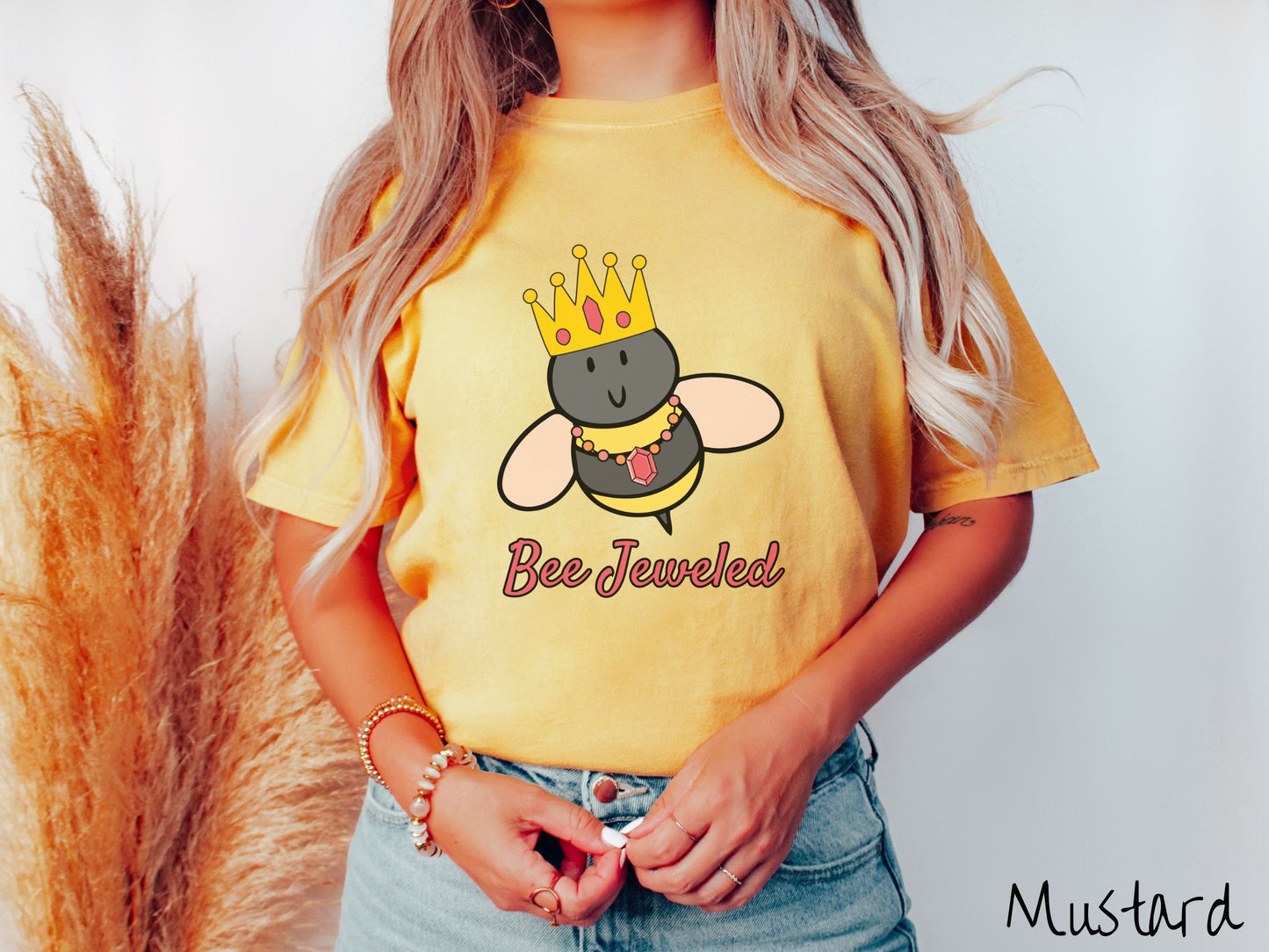 A woman wearing a cute, vintage mustard colored Comfort Colors T-shirt with the text Bee Jeweled in pink, cursive font. Above that is a black and yellow honey bee wearing a red, bejeweled golden crown, and a golden necklace with a large, red jewel.