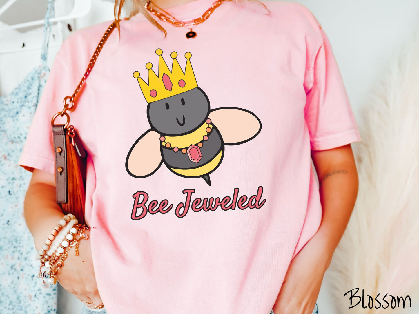A woman wearing a cute, vintage blossom colored Comfort Colors T-shirt with the text Bee Jeweled in pink, cursive font. Above that is a black and yellow honey bee wearing a red, bejeweled golden crown, and a golden necklace with a large, red jewel.