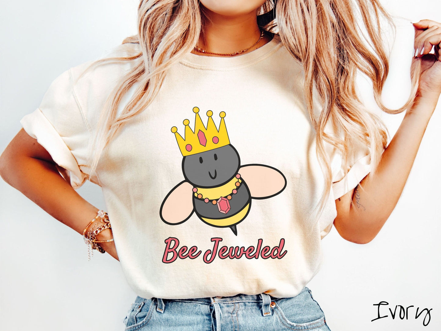 A woman wearing a cute, vintage ivory colored Comfort Colors T-shirt with the text Bee Jeweled in pink, cursive font. Above that is a black and yellow honey bee wearing a red, bejeweled golden crown, and a golden necklace with a large, red jewel.