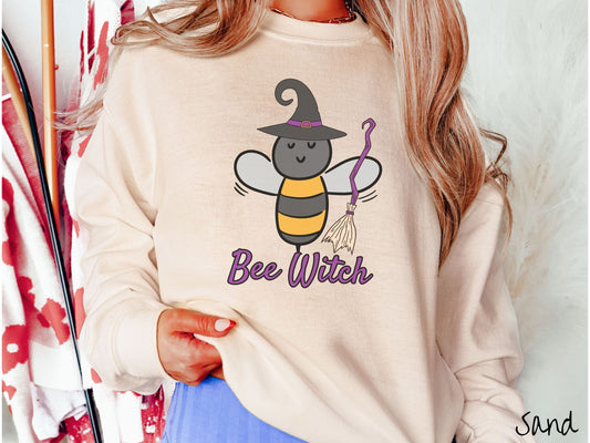 A woman wearing a cute, vintage sand colored comfy sweatshirt with the text Bee Witch in purple, cursive font. Above that is a black and yellow honey bee holding a purple, straw broom and wearing a black witch hat with a purple hat band.