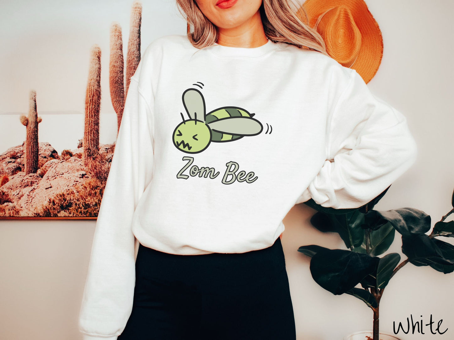 A woman wearing a cute, vintage white colored comfy sweatshirt with the text Zom Bee in light green, cursive font. Above that is a light and dark green honey bee with a crazy face buzzing in the air.