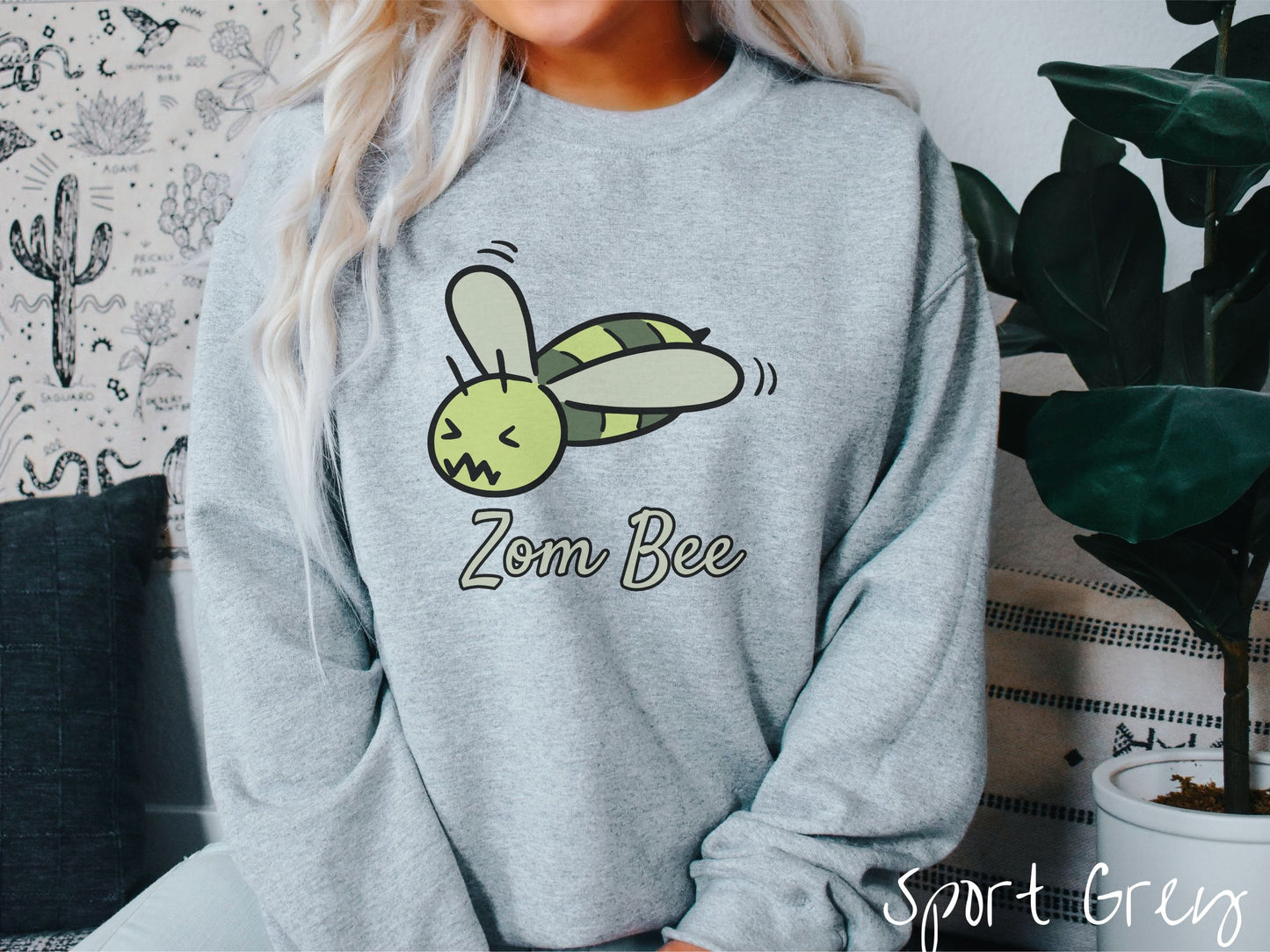 A woman wearing a cute, vintage sport grey colored comfy sweatshirt with the text Zom Bee in light green, cursive font. Above that is a light and dark green honey bee with a crazy face buzzing in the air.