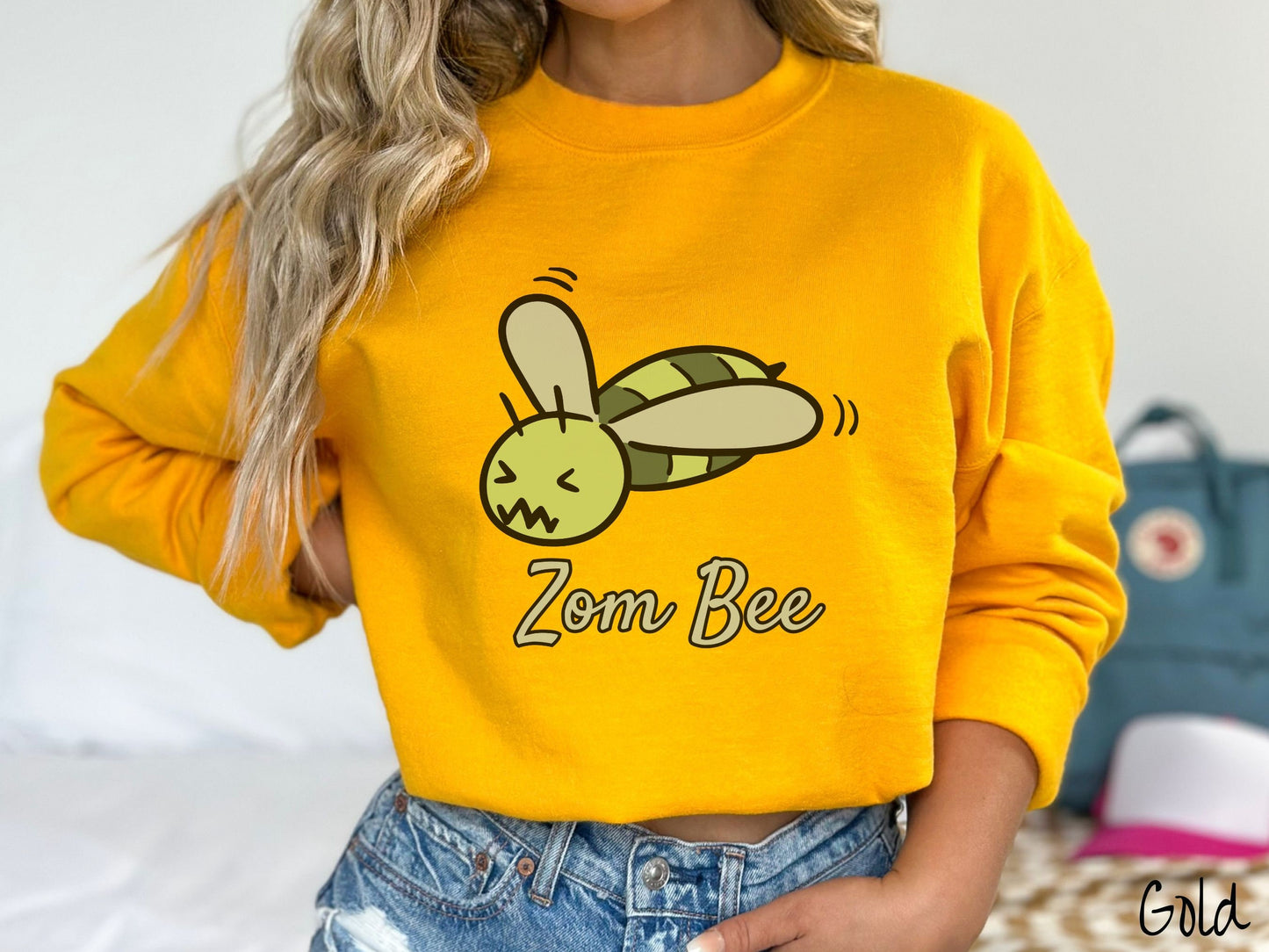 A woman wearing a cute, vintage gold colored comfy sweatshirt with the text Zom Bee in light green, cursive font. Above that is a light and dark green honey bee with a crazy face buzzing in the air.