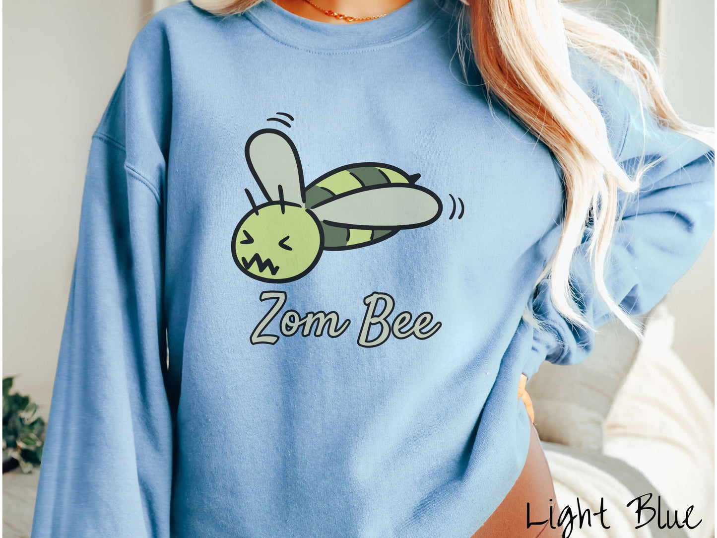 A woman wearing a cute, vintage light blue colored comfy sweatshirt with the text Zom Bee in light green, cursive font. Above that is a light and dark green honey bee with a crazy face buzzing in the air.