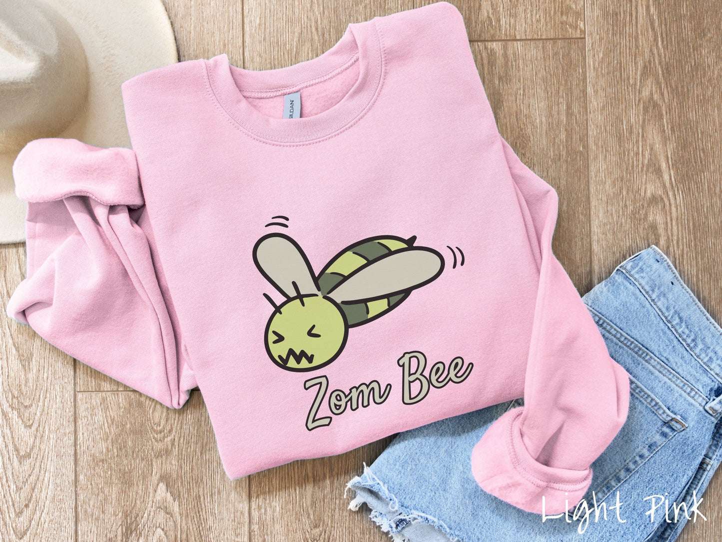 A cute, vintage light pink colored comfy sweatshirt with the text Zom Bee in light green, cursive font. Above that is a light and dark green honey bee with a crazy face buzzing in the air.
