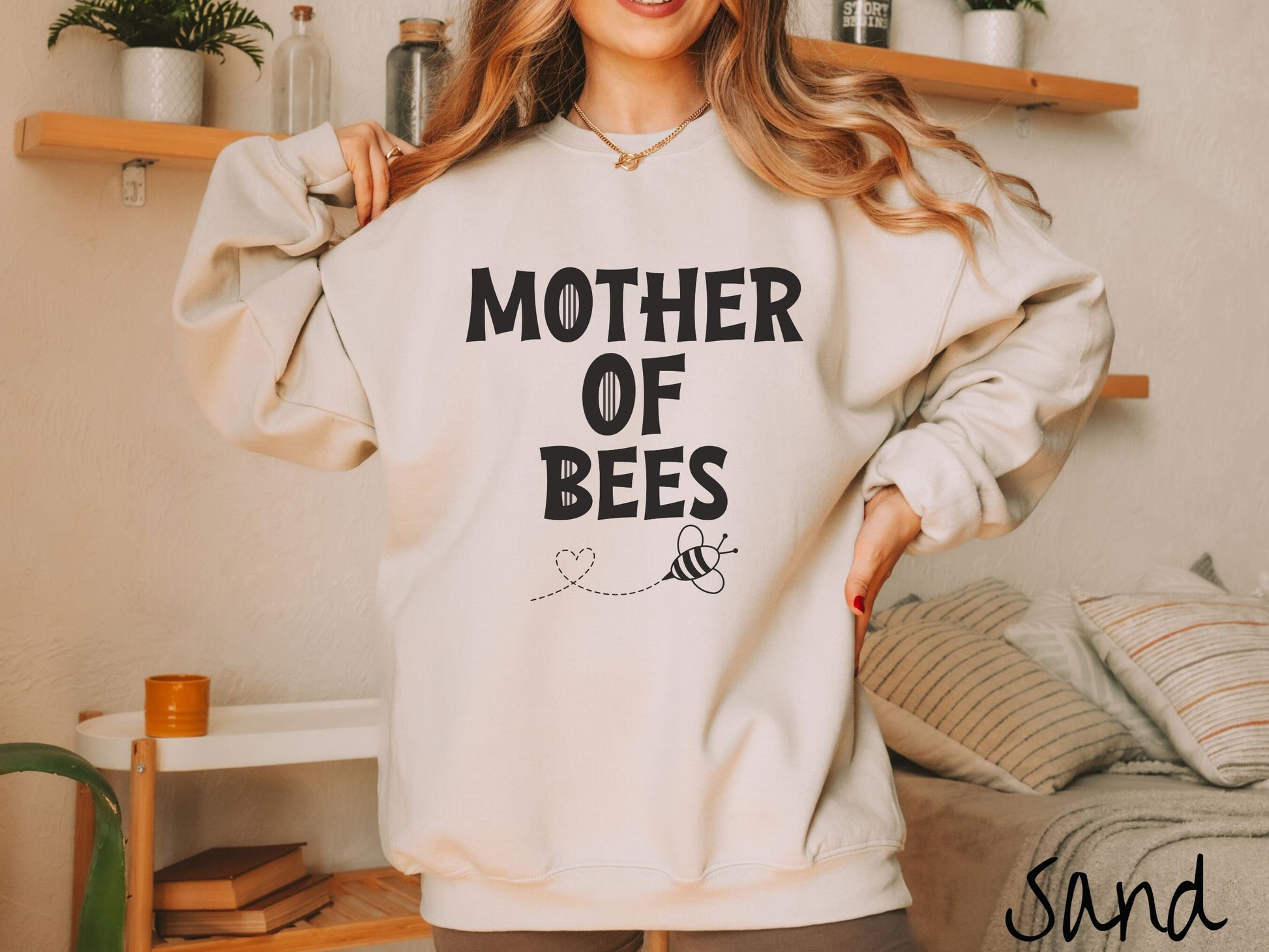 A woman wearing a cute, vintage sand colored comfy sweatshirt with the text Mother of Bees in black, uppercase font. Below that is a black outlined bee buzzing around, the trail it leaves has created a heart shape in the air.