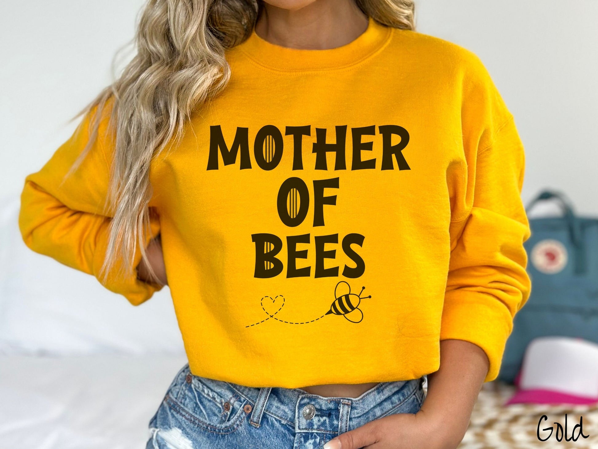 A woman wearing a cute, vintage gold colored comfy sweatshirt with the text Mother of Bees in black, uppercase font. Below that is a black outlined bee buzzing around, the trail it leaves has created a heart shape in the air.