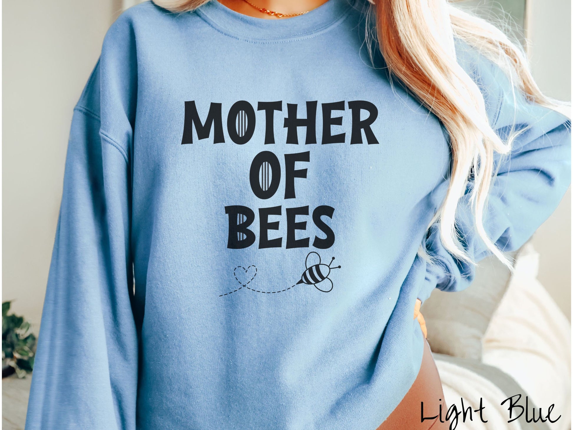 A woman wearing a cute, vintage light blue colored comfy sweatshirt with the text Mother of Bees in black, uppercase font. Below that is a black outlined bee buzzing around, the trail it leaves has created a heart shape in the air.