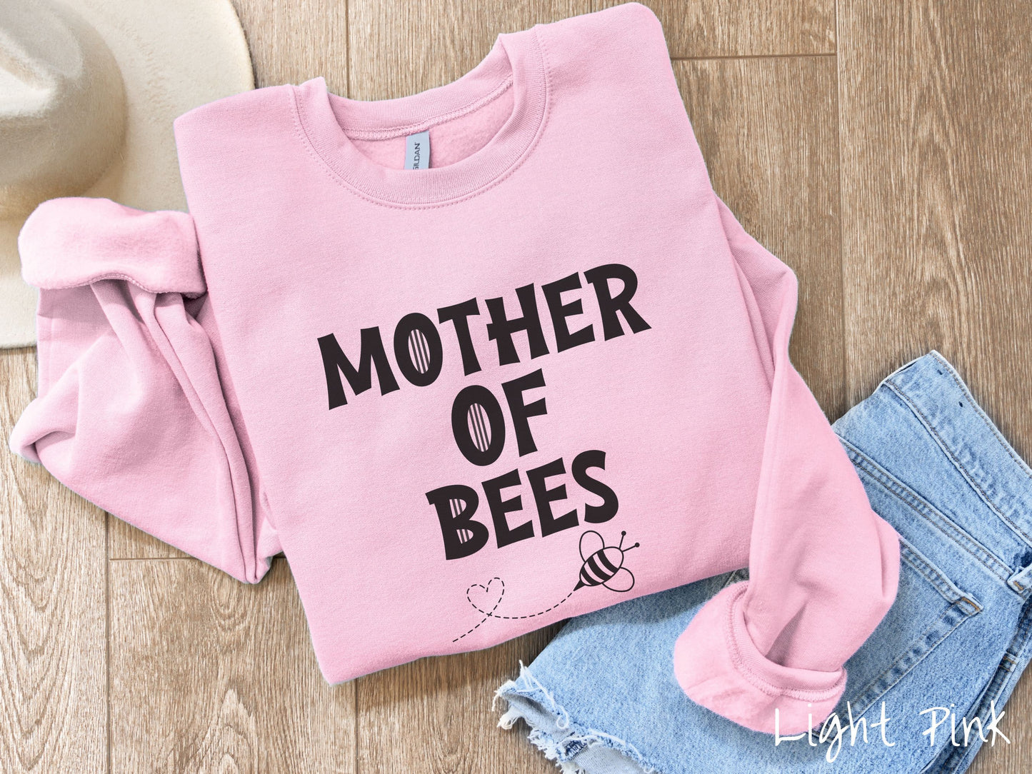 A cute, vintage light pink colored comfy sweatshirt with the text Mother of Bees in black, uppercase font. Below that is a black outlined bee buzzing around, the trail it leaves has created a heart shape in the air.