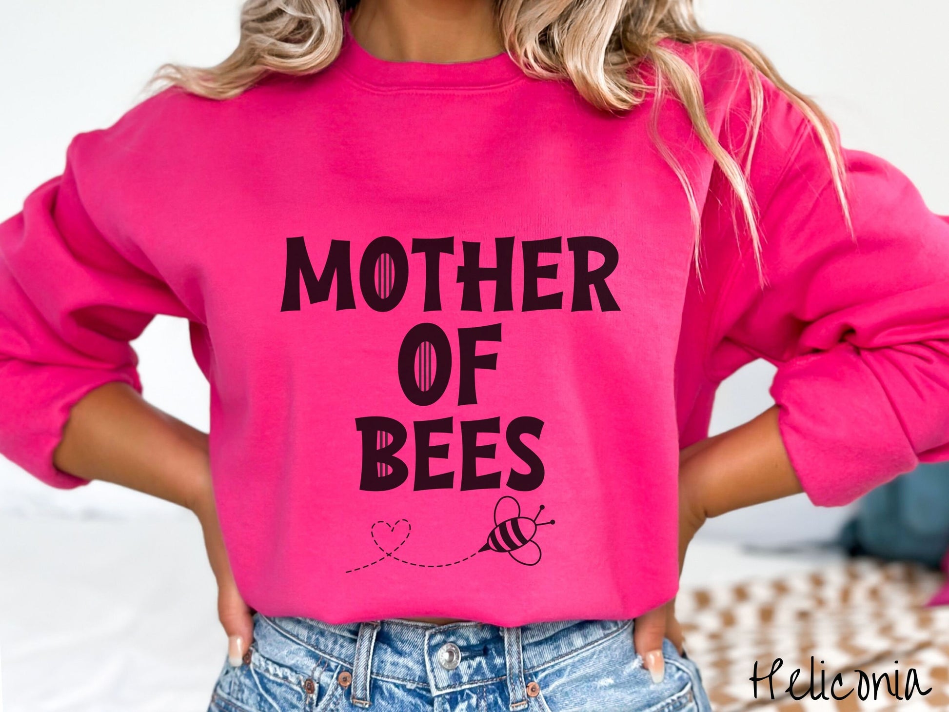 A woman wearing a cute, vintage heliconia colored comfy sweatshirt with the text Mother of Bees in black, uppercase font. Below that is a black outlined bee buzzing around, the trail it leaves has created a heart shape in the air.