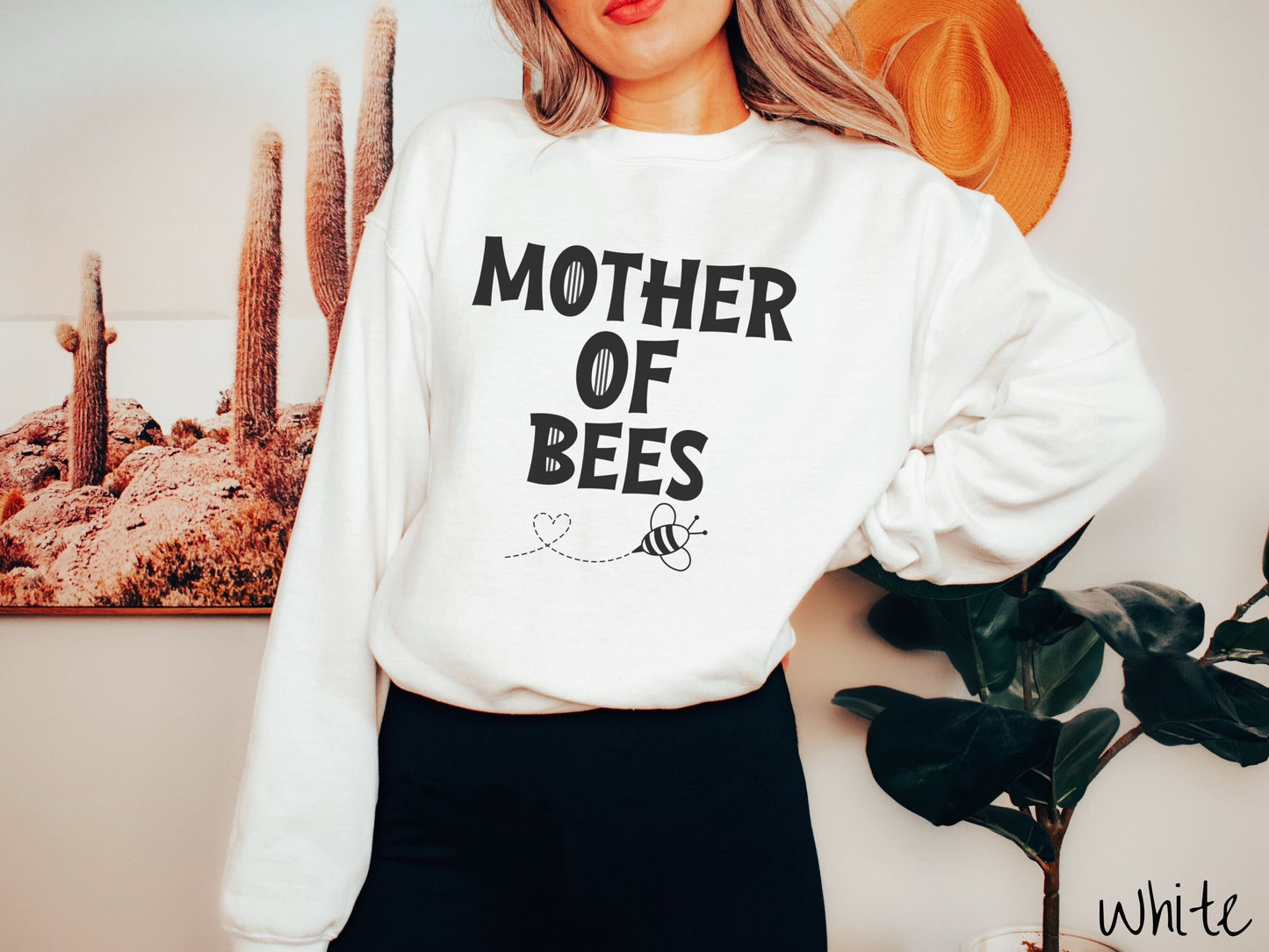 A woman wearing a cute, vintage white colored comfy sweatshirt with the text Mother of Bees in black, uppercase font. Below that is a black outlined bee buzzing around, the trail it leaves has created a heart shape in the air.