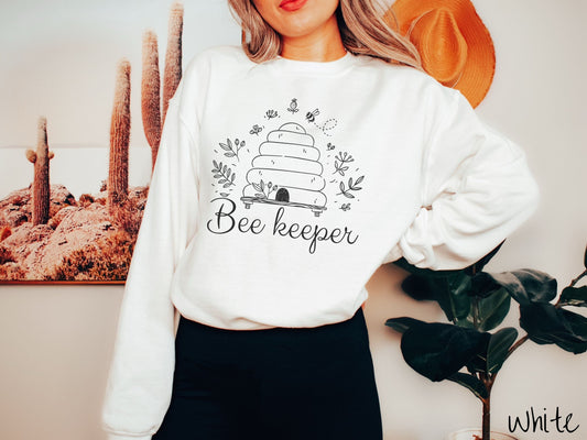 A woman wearing a cute, vintage white colored comfy sweatshirt with the text Beekeeper across the front in black, cursive font. Above this is a black-outlined bee hive with bees buzzing around it and there are leaves in the air.