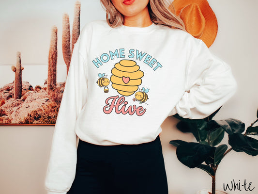 A woman wearing a cute, vintage white colored comfy sweatshirt with the text Home Sweet Hive in cyan and red font. In between the words are a yellow bee hive with a red heart on it and smiling honey bees flying around holding flowers.