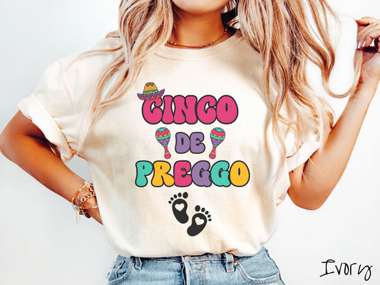 A woman wearing a cute, vintage ivory colored Comfort Colors t-shirt with the text Cinco de Preggo in colorful font. A sombrero is hanging on top of the C in Cinco, colorful maracas around the word de, and two baby footprints with hearts inside.