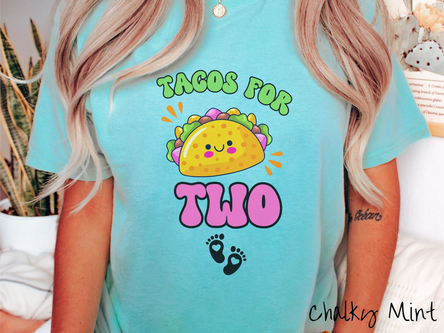 A woman wearing a cute, vintage chalky mint colored Comfort Colors t-shirt with text Tacos for Two in green and pink font. In between the font is a smiling taco with colorful fillings and below are two baby footprints with hearts inside.