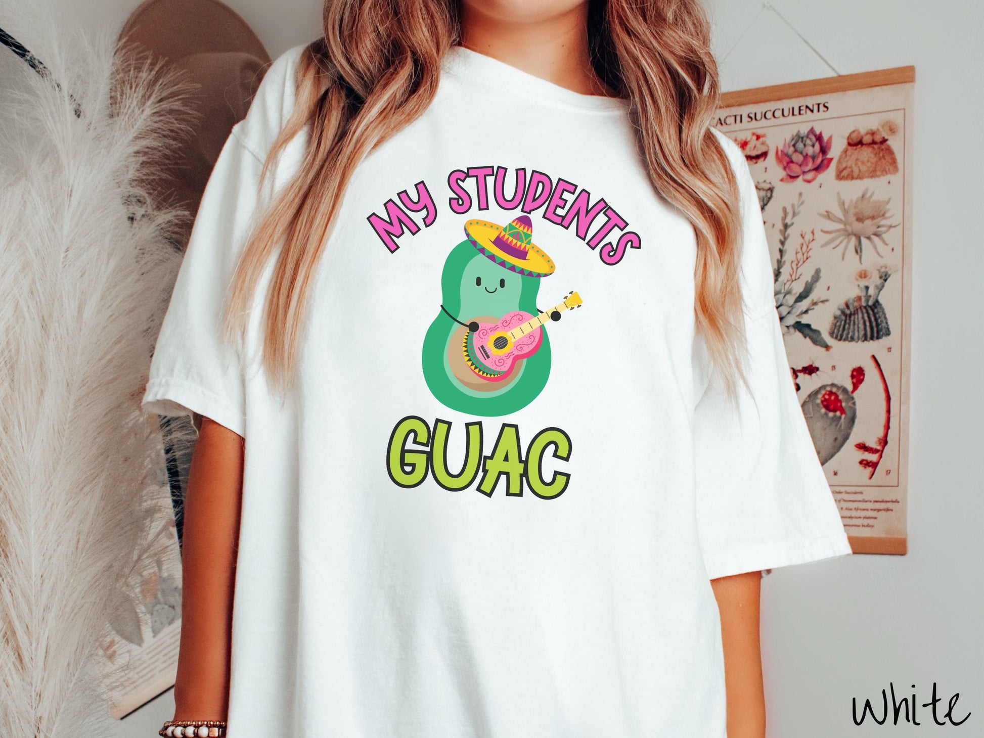 A woman wearing a cute, vintage white colored Comfort Colors t-shirt with the text My Students Guac in pink and green font. In between the text is a smiling avocado wearing a colorful sombrero and playing a colorful mariachi guitar.