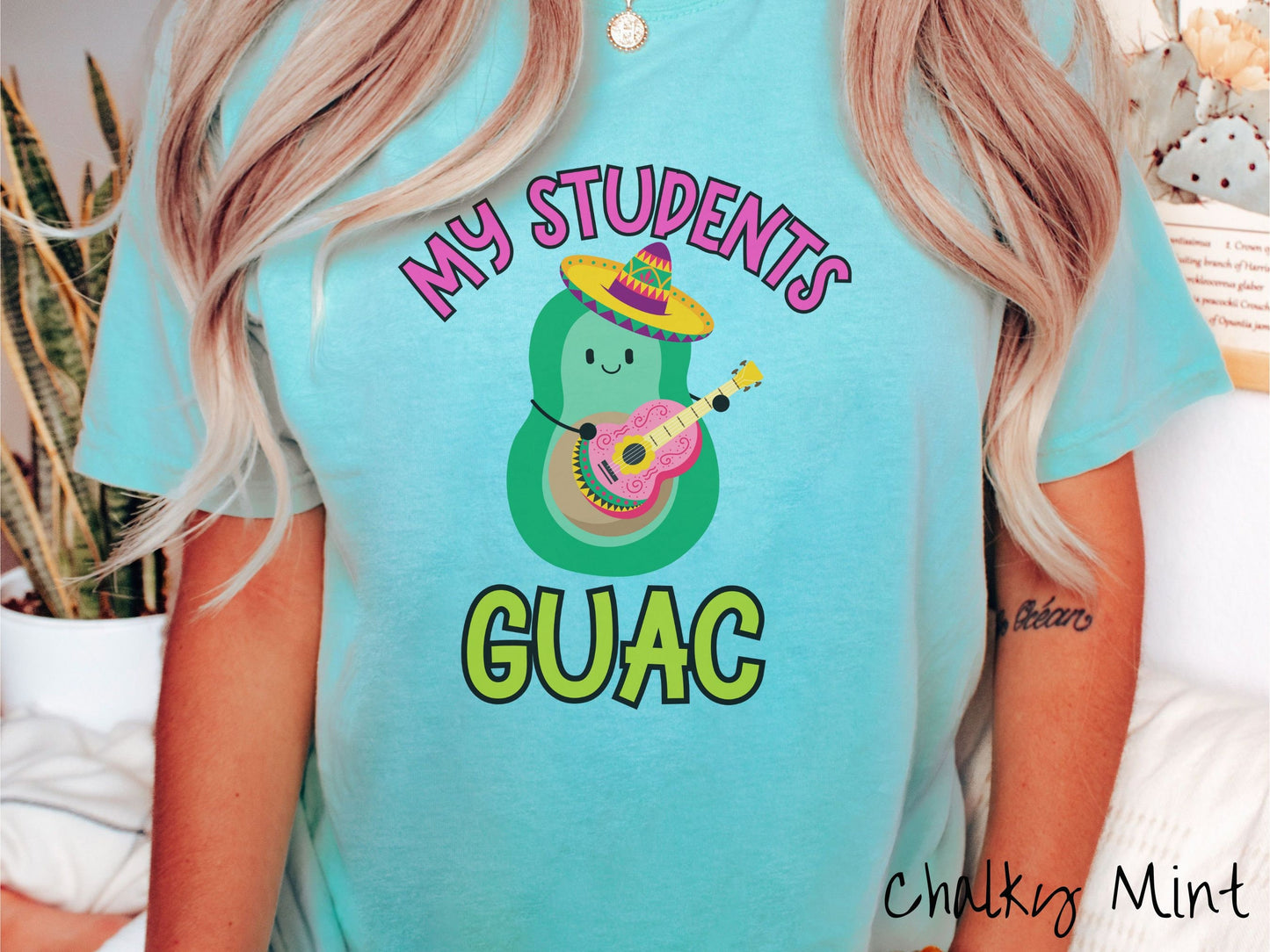 A woman wearing a cute, vintage chalky mint colored Comfort Colors t-shirt with the text My Students Guac in pink and green font. In between the text is a smiling avocado wearing a colorful sombrero and playing a colorful mariachi guitar.