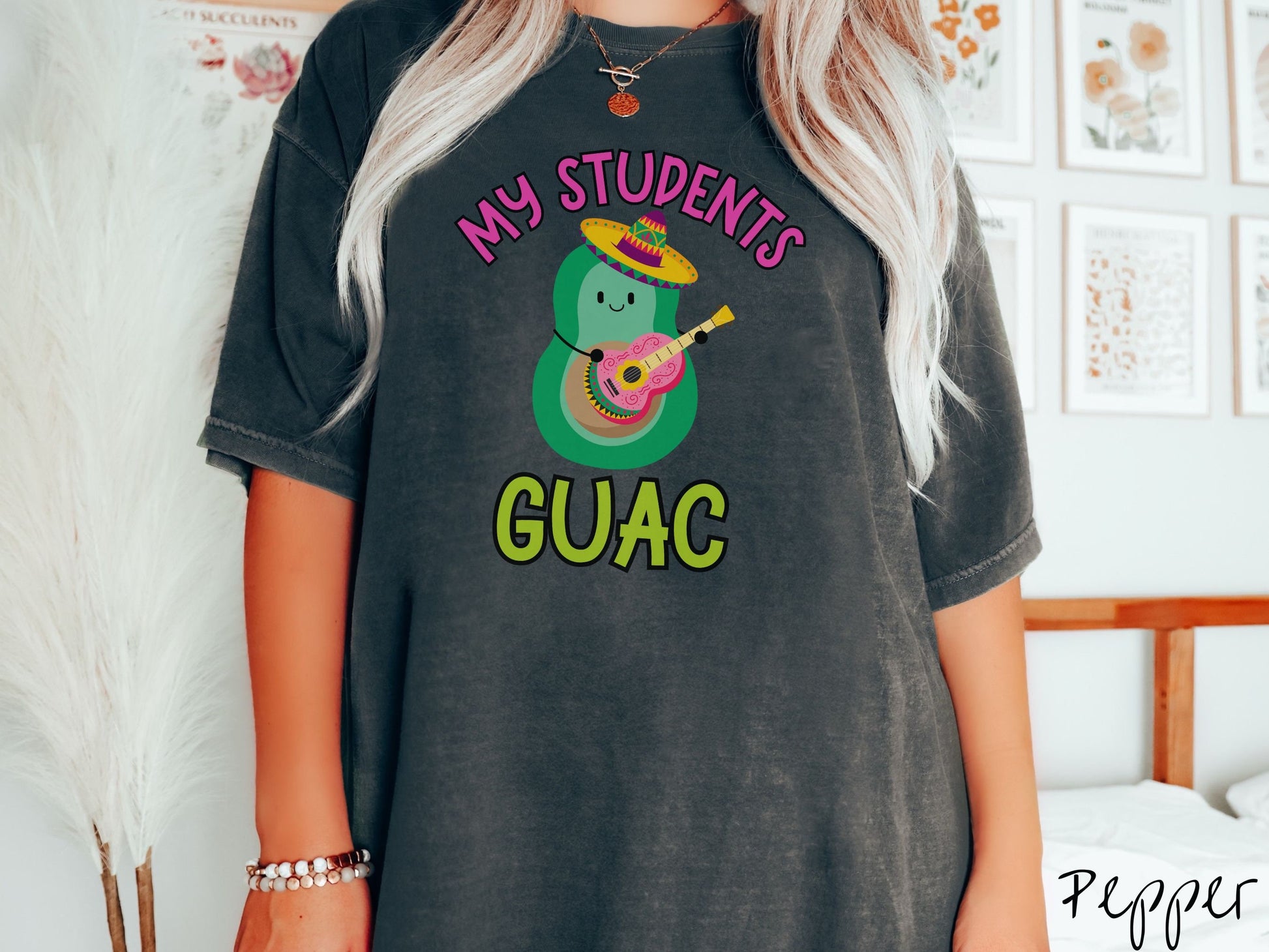 A woman wearing a cute, vintage pepper colored Comfort Colors t-shirt with the text My Students Guac in pink and green font. In between the text is a smiling avocado wearing a colorful sombrero and playing a colorful mariachi guitar.