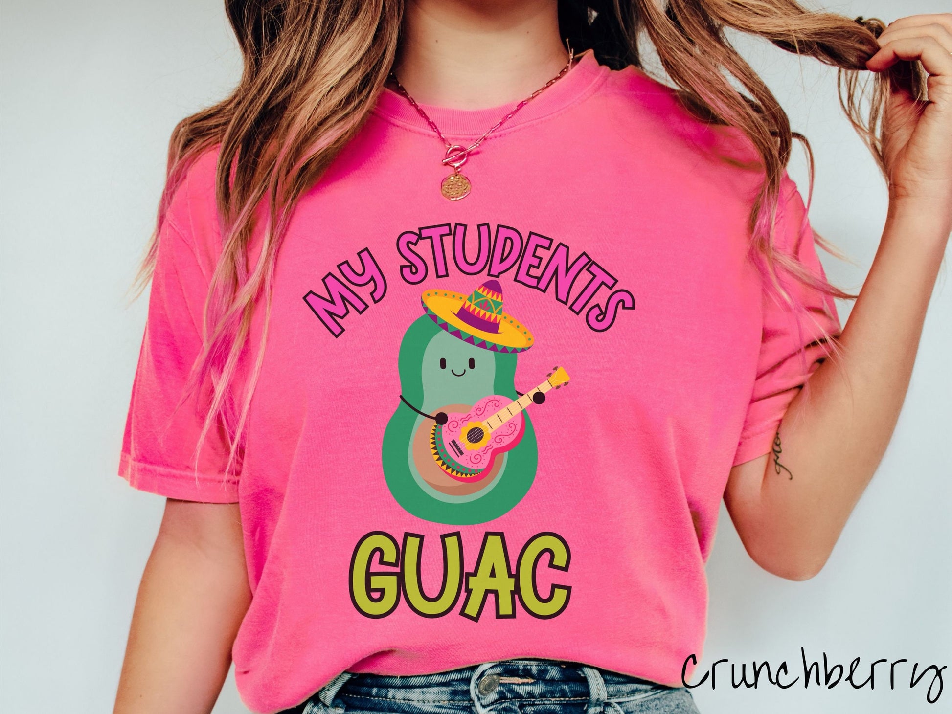 A woman wearing a cute, vintage crunchberry colored Comfort Colors t-shirt with the text My Students Guac in pink and green font. In between the text is a smiling avocado wearing a colorful sombrero and playing a colorful mariachi guitar.